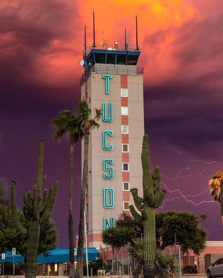 A tall building with the word 'TUCSON' on its side, surrounded by cacti and palm trees under a dramatic sky with lightning. 2