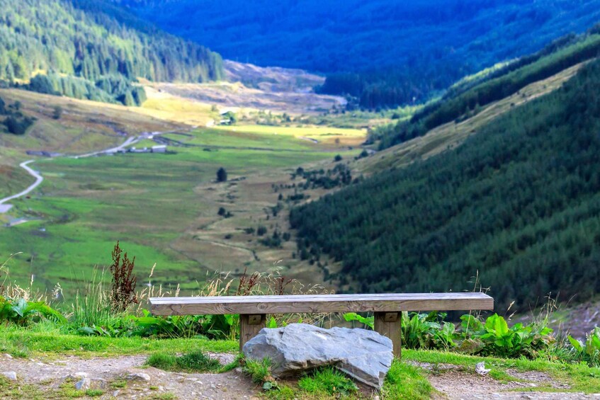 A wooden bench on a grassy hill overlooking a valley with a winding road and forested hills. 2