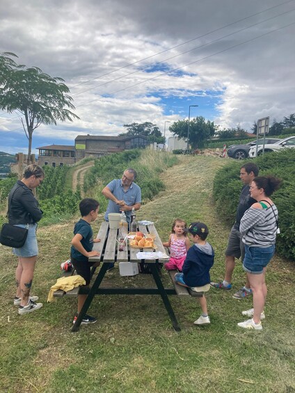 A group of people around a picnic table with food and drinks, set in a grassy outdoor area with buildings in the background. 1