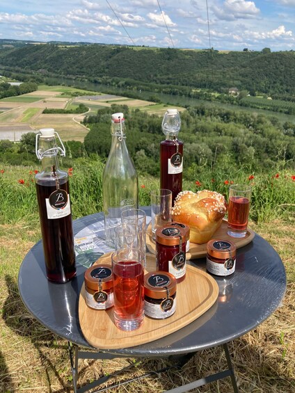 A picnic setup with a bottle of Brumetot wine, a glass of red wine, a bottle of rosé, a jar of jam, a loaf of bread, and a glass of water on a table. 3