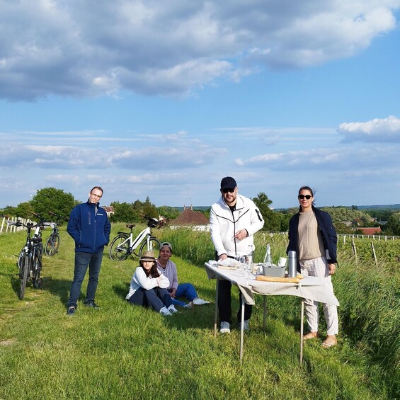 A group of people outdoors with bicycles, a picnic table, and a rural landscape in the background. 2