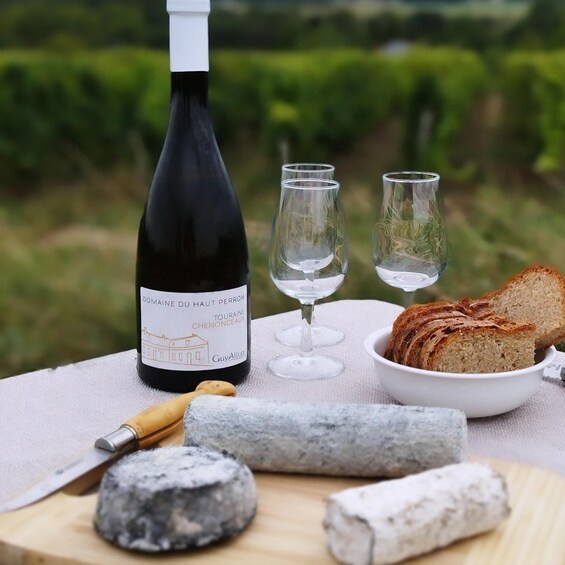 A bottle of Domaine du Haut Perron wine, two glasses, a bowl of bread, and a cheese wheel on a table outdoors. 1