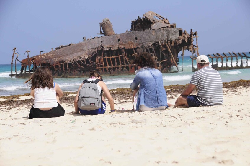 A group of people sitting on a beach, looking at a rusted shipwreck in the ocean. 1