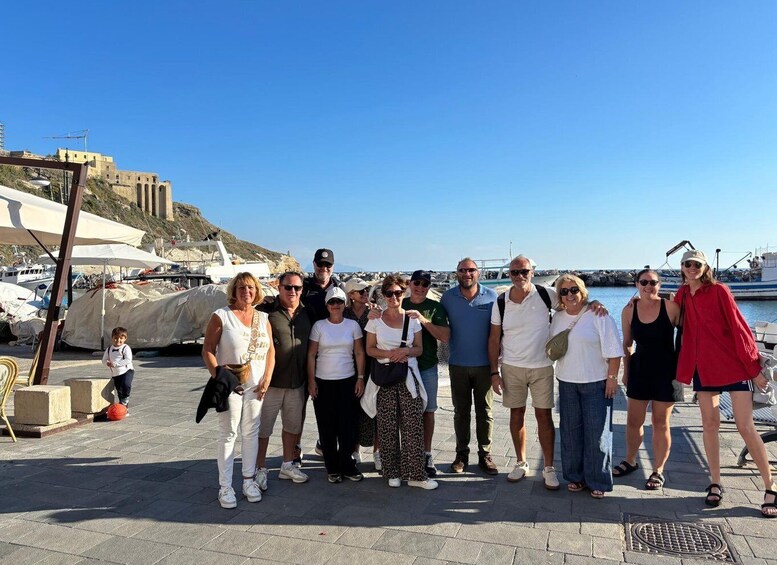 A group of people standing on a paved area near a body of water with boats and a historic building in the background. 4