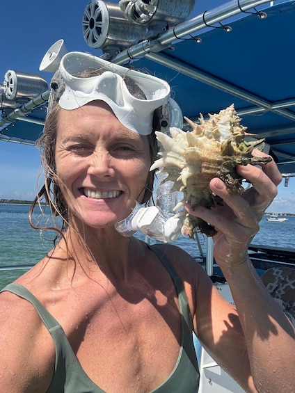 A person holding a large shell, wearing a snorkel mask, on a boat with a clear blue sky and calm sea in the background. 1