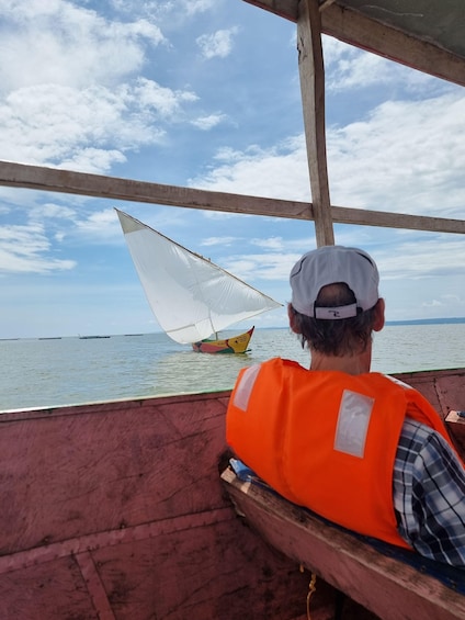 A person in an orange vest and white cap on a boat, looking at a sailboat with a colorful hull and a white sail. 1
