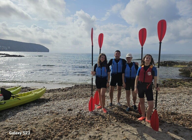 Four people on a rocky beach holding paddles, with a green kayak and a body of water in the background. 4