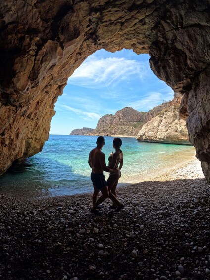 A couple standing in a rocky cave opening with a beach and clear blue sea in the background. 5