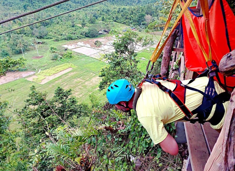 A person rappelling down a rope with a helmet and harness, overlooking a lush green landscape with terraced fields. 4