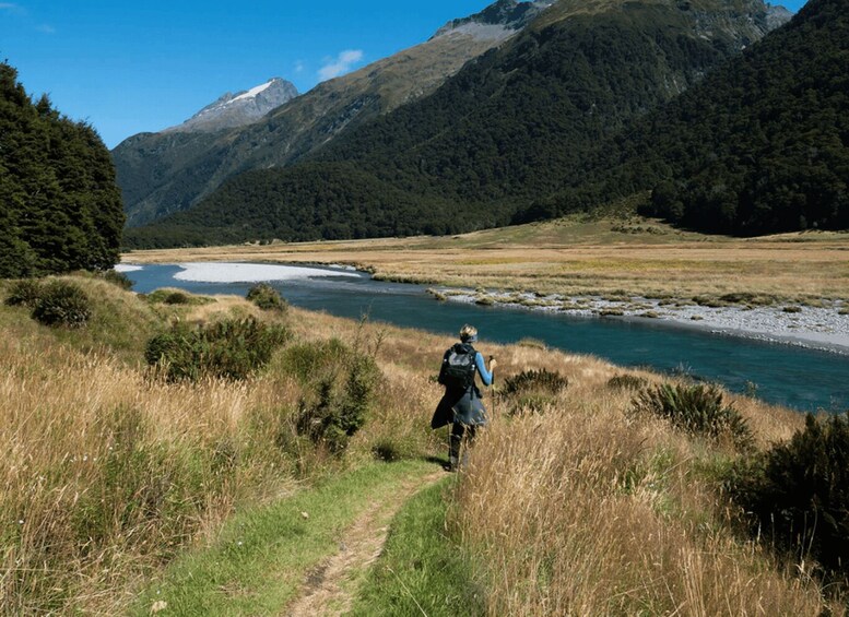 A hiker walking along a trail beside a river with mountains in the background. 1