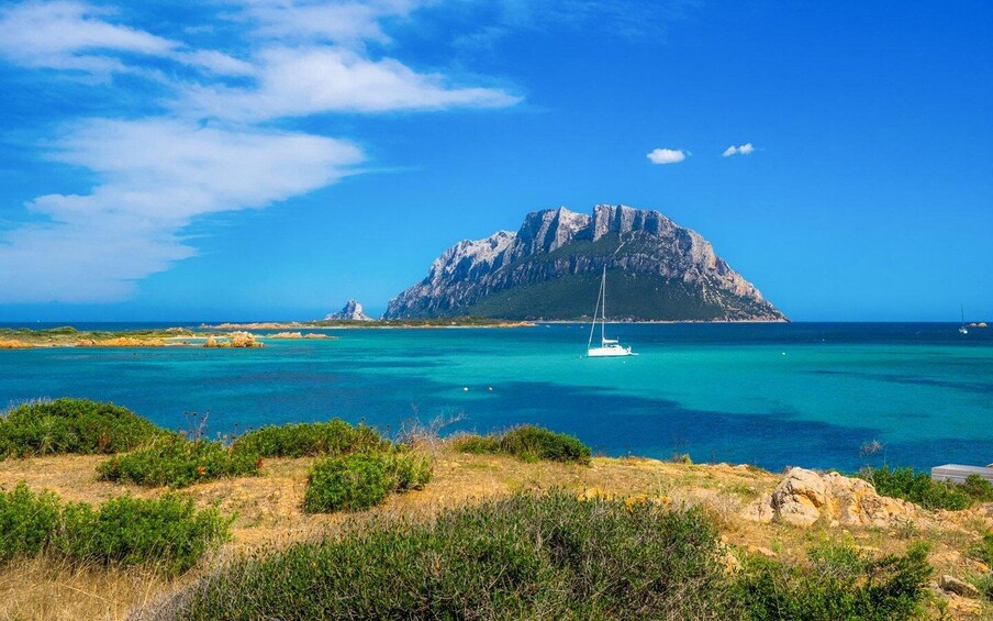 A coastal landscape with a prominent rocky mountain, clear turquoise waters, and a sailboat. 1