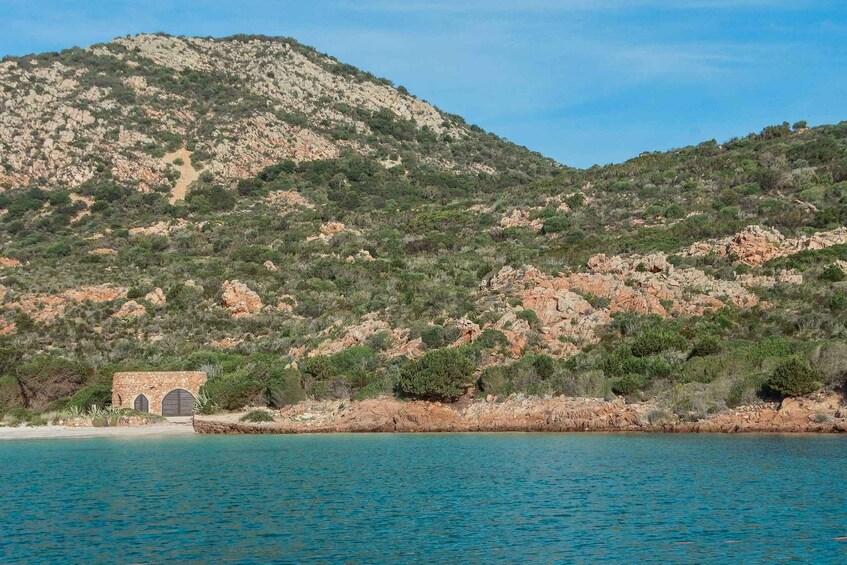 A coastal landscape with a rocky hill, a small stone building, and clear blue waters. 4