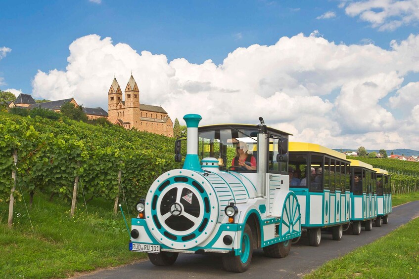 A white and turquoise train with passengers, traveling on a road with vineyards and a historic building in the background. 2