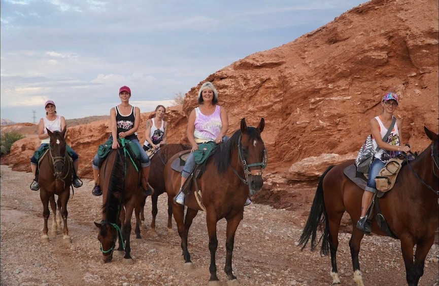 Four people riding horses on a dirt path with red rock formations in the background. 3