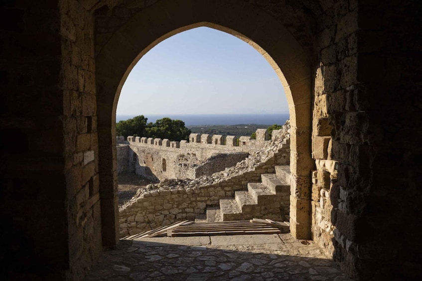 An ancient stone archway leading to a fortified wall with a view of the sea and distant landscape. 1