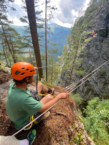 A person sitting on a rope bridge in a forested area with a mountain in the background. 2