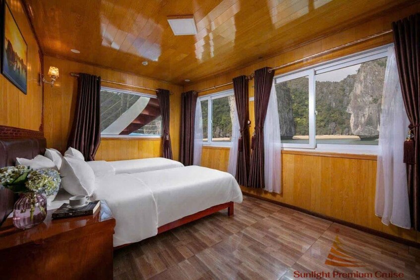 A cabin with wooden walls and floors, two beds, a nightstand, a lamp, and large windows with curtains. 1