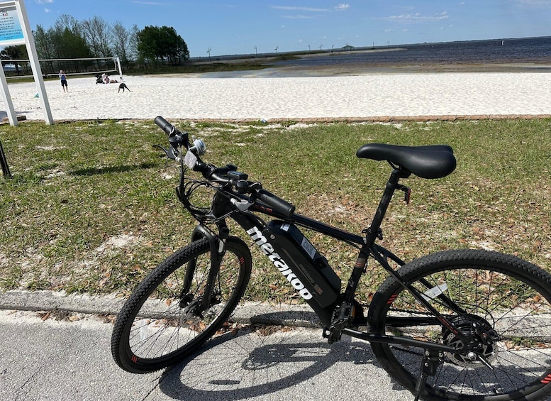 A black bicycle with a water bottle and a black seat parked on a paved area near a sandy beach. 1