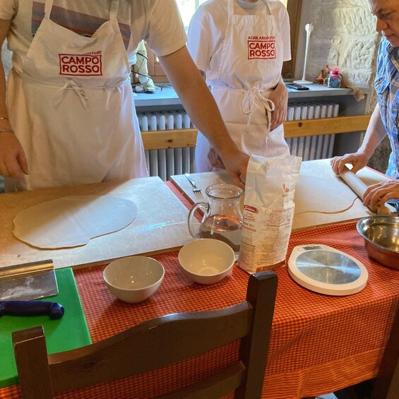 Two people wearing white aprons with 'CAMPO ROSSA' logos, preparing food on a table with various kitchen items like bowls, a rolling pin, and a bottle of sauce. 2
