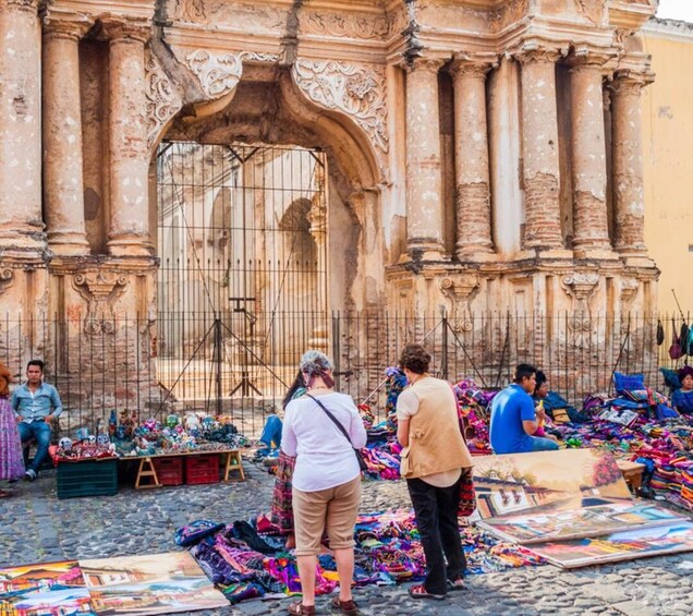 A street market with colorful textiles and clothing stalls in front of a historic building with ornate columns. 5