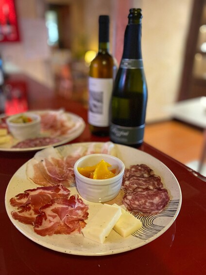 A plate of cured meats, cheese, and a small bowl of fruit on a table with bottles of wine in the background. 4