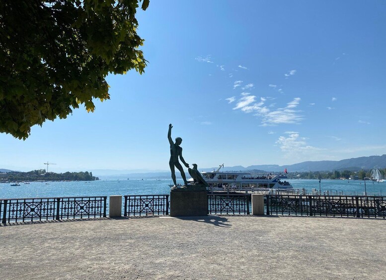 A statue of a man with raised arms on a platform by the waterfront, with boats and a distant island in the background. 2