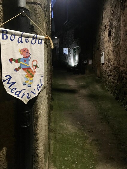A narrow alleyway with a banner hanging on a wall, a stone building in the background, and a sign with text. 5