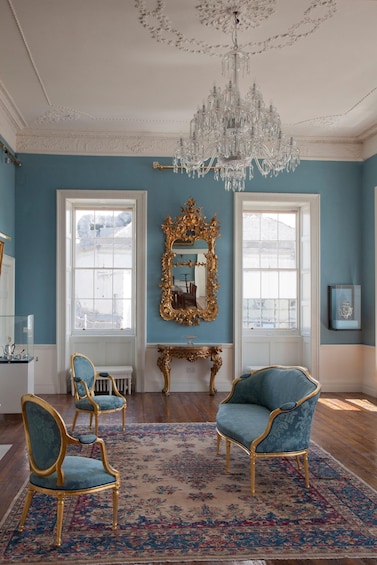 A room with blue walls, a chandelier, two blue armchairs, a gold-framed mirror, and a floral carpet. 1