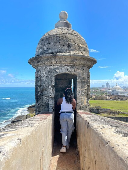 A person entering a stone structure with a dome, overlooking a coastal area with buildings and a clear sky. 1