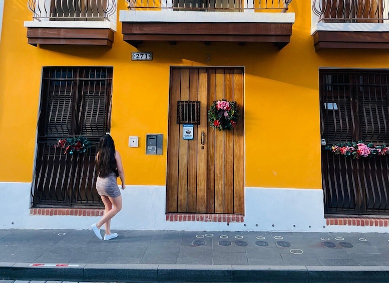 A person walking past a yellow building with a wooden door, a number plate, and a wreath on the door. 5