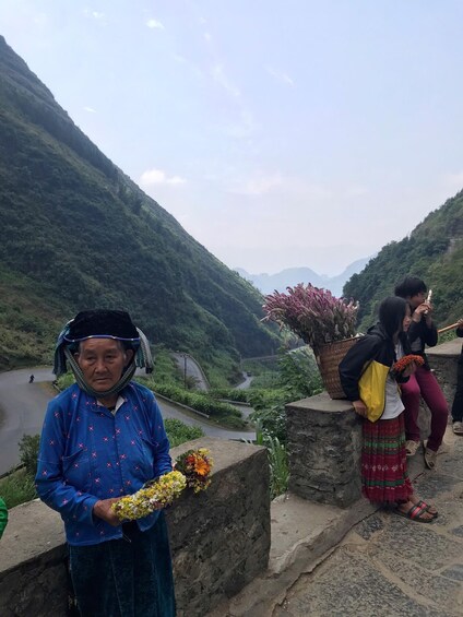 A mountainous landscape with a river, a person holding flowers, and another person sitting on a ledge. 2