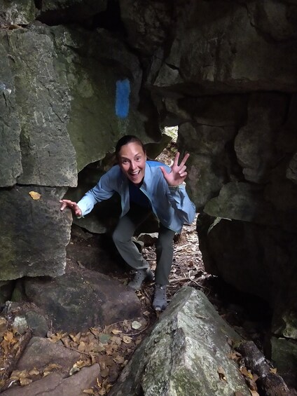 A person crouching in a rocky cave with moss-covered walls and scattered leaves on the ground. 5