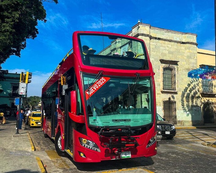 A red double-decker bus with a 'Nuevo 2025' sign, parked on a street with buildings and a clear blue sky in the background. 1