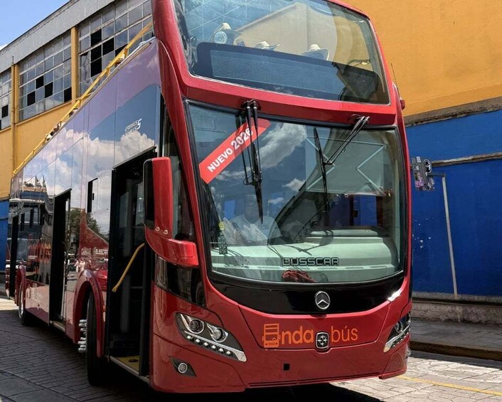A red double-decker bus with 'Busscar' and 'anda bus' branding, parked in front of a building with a blue wall. 2