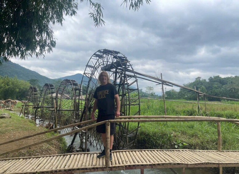 A person standing on a bamboo bridge over a water channel with traditional water wheels in the background. 2