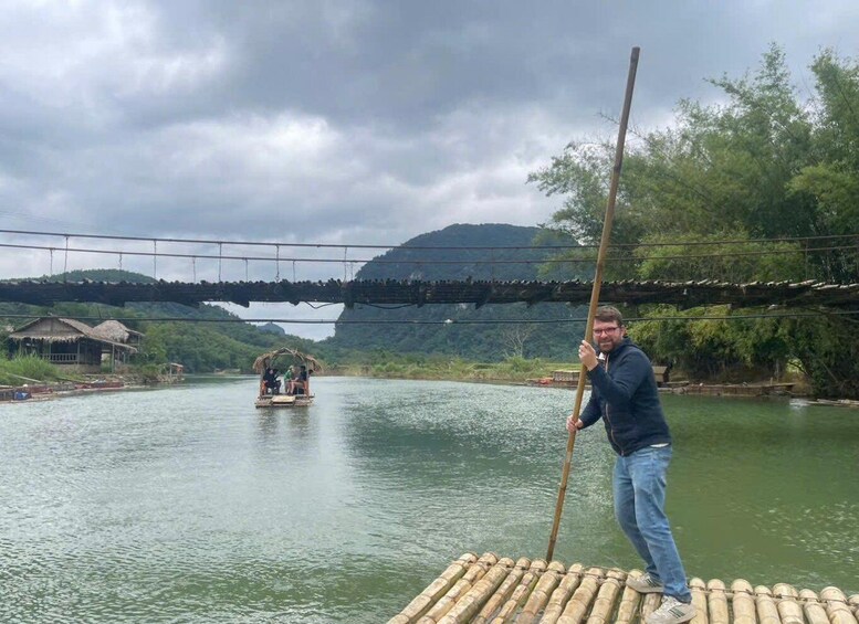 A person standing on a bamboo raft holding a long pole, with a small boat in the background on a river surrounded by greenery and a bridge overhead. 5