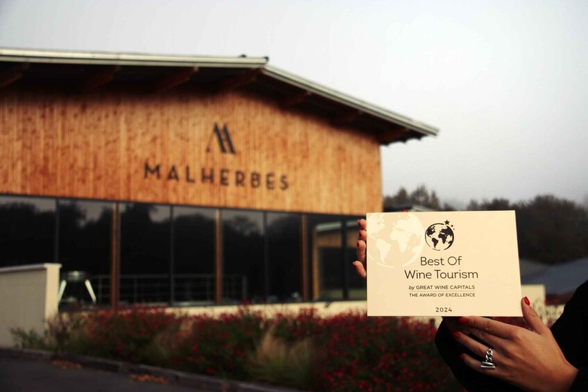 A person holding a certificate titled 'Best Of Wine Tourism' in front of a building with the sign 'MALHERBES'. 3