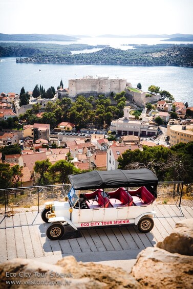 A tour vehicle on a paved path overlooking a coastal town with a historic castle and a body of water in the background. 4