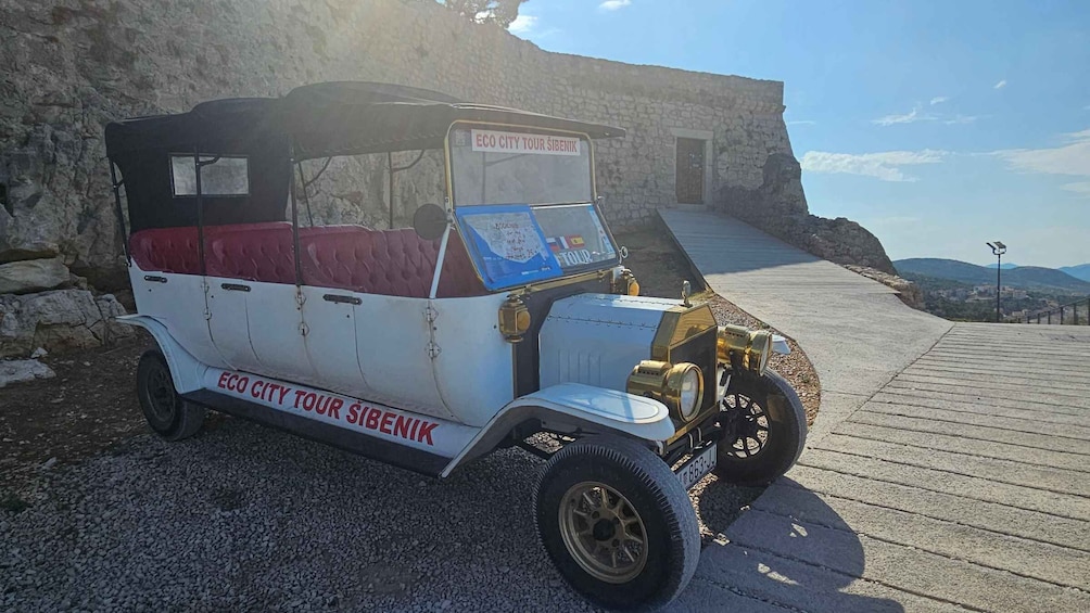 A vintage car with 'ECO CITY TOUR SIBENIK' written on it, parked on a gravel path with a stone wall and a clear sky in the background. 1