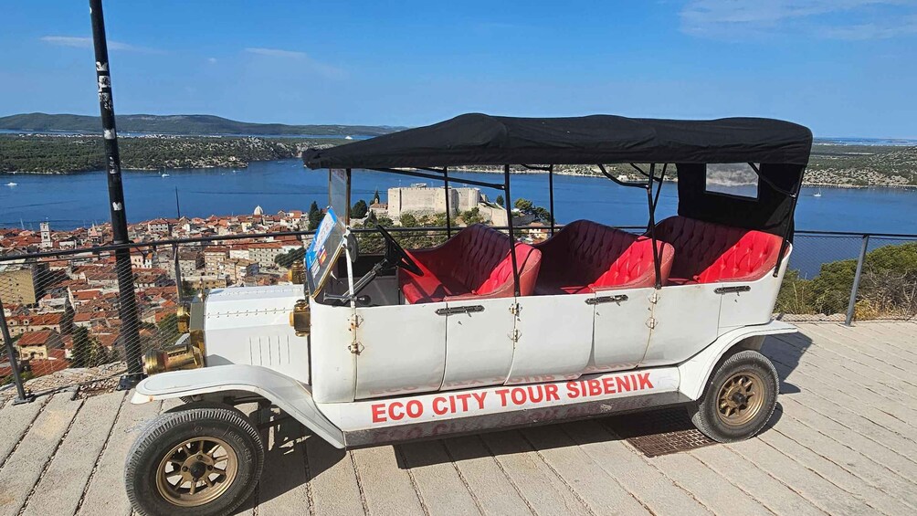 A white tour vehicle with red seats and a black canopy parked on a paved area with a scenic view of a coastal town and water. 3