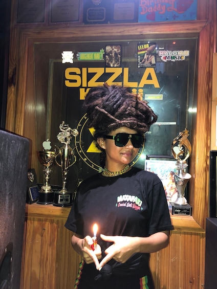 A person holding a lighter with a flame, in front of a wall with various awards and a sign that reads 'SIZZLA'. 4