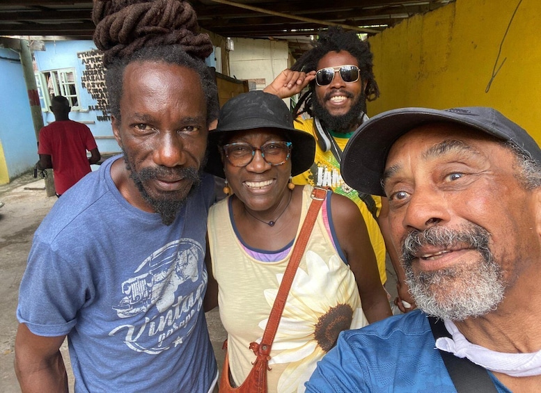 Four people standing close together in an outdoor setting with a yellow wall and a blue building in the background. 1