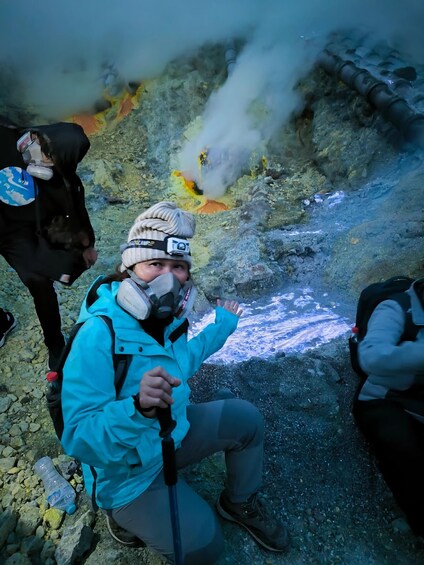 A person in a blue jacket and beanie kneeling on rocky terrain with a water bottle and a walking stick. 3