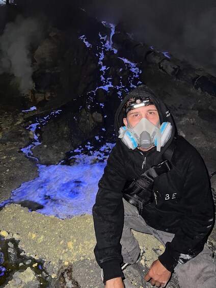 A person in a black jacket with a helmet and breathing apparatus, standing on rocky terrain with blue glowing ice formations. 5