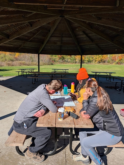 A group of people sitting at a picnic table under a wooden pavilion in a park with autumn foliage in the background. 3