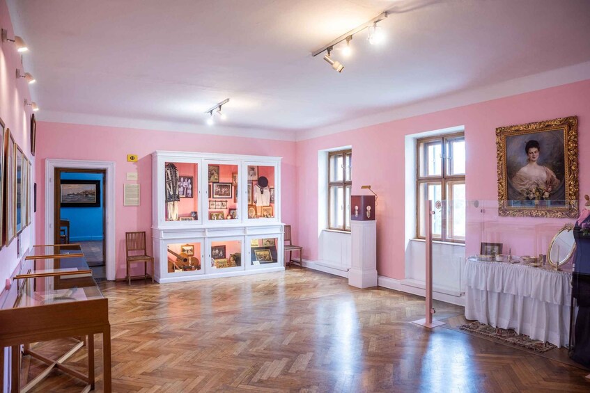 A pink-walled room with wooden floors, a white cabinet with glass doors, and a framed portrait. 1