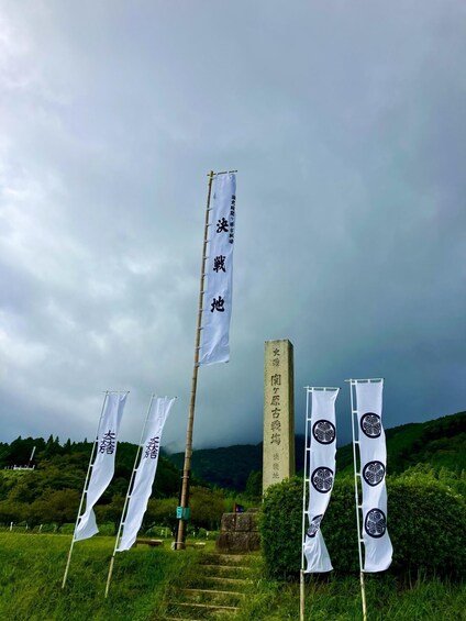 A series of white flags with black text, a stone monument, and a grassy landscape under a cloudy sky. 2