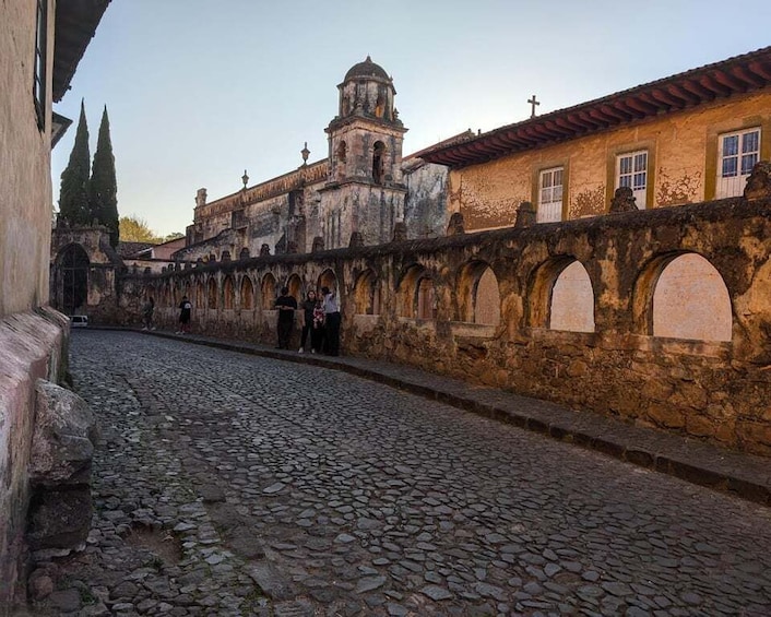 A cobblestone street with historic buildings, a church with a bell tower, and arches along the walkway. 1