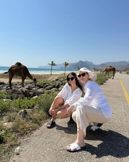Two people posing on a roadside with a beach and camels in the background. 1