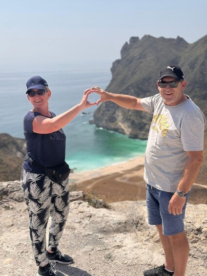 Two people making a heart shape with their hands, standing on a rocky cliff with a beach and ocean in the background. 5